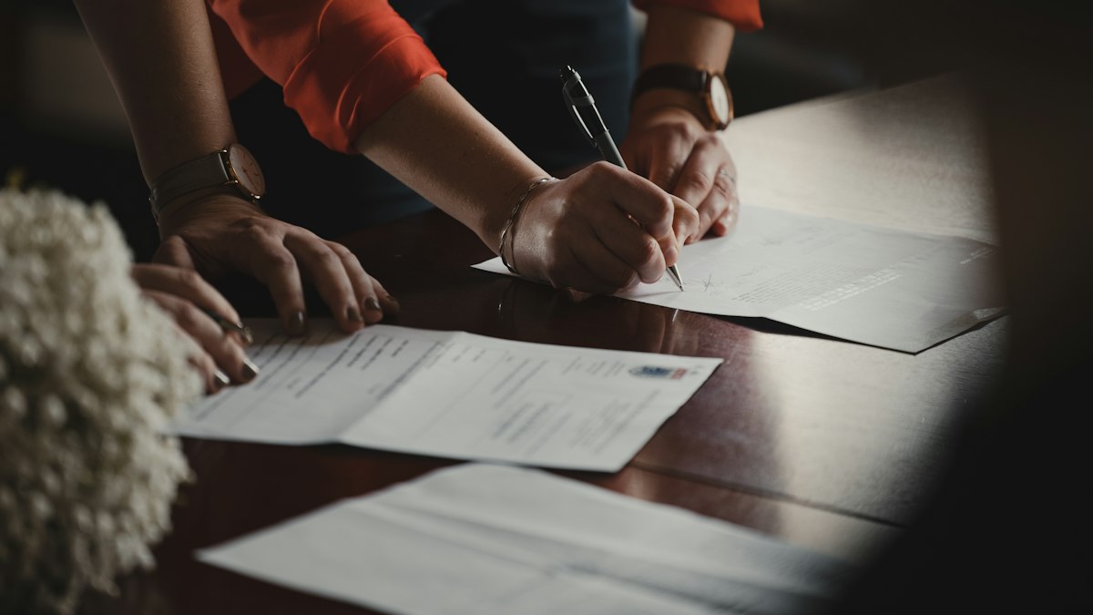 Deux personnes signent des documents sur une table en bois sombre, l'une tient un stylo au-dessus d'un contrat